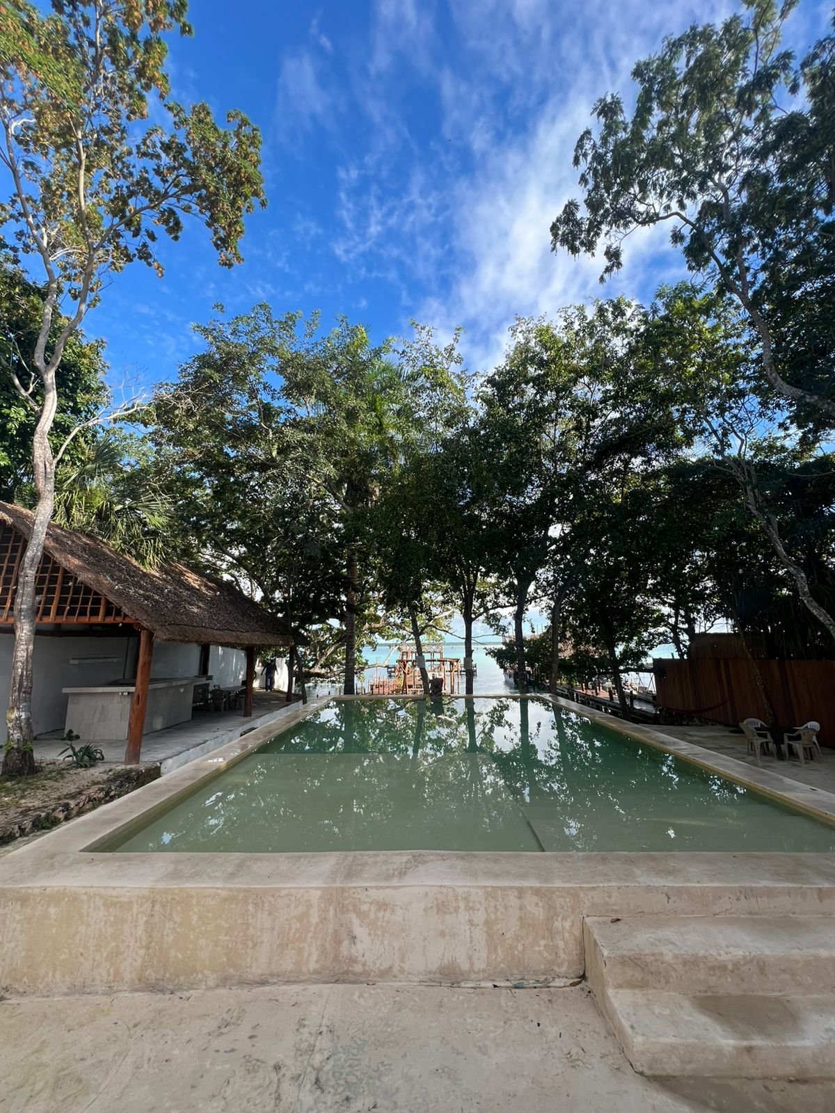 Alberca del Hotel Casa del Ángel Bacalar rodeada de árboles y palapa con la laguna al fondo
